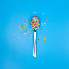 Sprinkles in a spoon, flat lay, blue background, square image. Baking or holiday concept