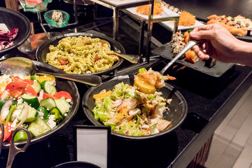 Hand holding a silver stainless spoon with mixed fruit and seafood salad over the salad bowl in buffet line