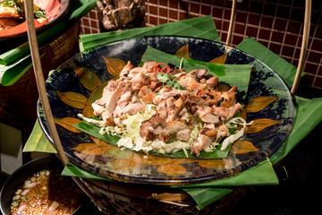 Charcoal grilled marinated chicken slices in Thai style arranged on banana leaves in ceramic hand painted flat dish over traditional basket.