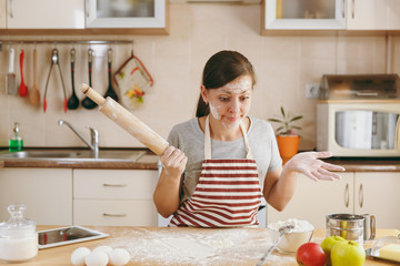 A young perplexed and confused woman sitting at a table with rolling pin and flour and going to...