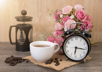 Hot cup of coffee and alarm clock on wood table with rose flower.