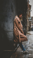 Lifestyle full portrait of young attractive fashionable Caucasian female in brown coat standing in the narrow street in Europe. Vertical photo