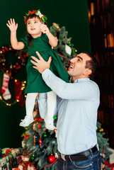 Dad holds his little daughter in green dress posing before a Christmas tree