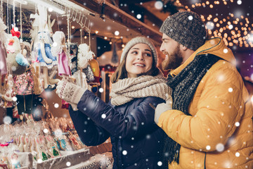 Happy young attractive lady is choosing the gift in a souvenir shop outdoors on a fairy, her lover is embracing her, smiling, they dressed in winter outfits, hats, snowfall