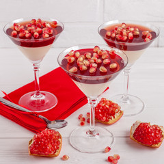 Creamy vanilla panna cotta with red jelly in beautiful glasses, fresh ripe pomegranate on white wooden background.