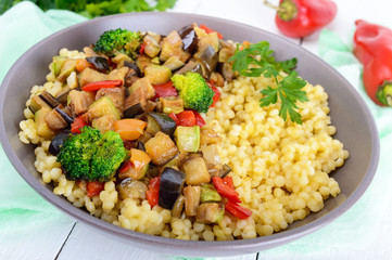 Warm vegan salad with couscous and fried vegetables (eggplant, zucchini, onion, sweet pepper, broccoli) in a ceramic bowl on a white wooden background. Proper nutrition.