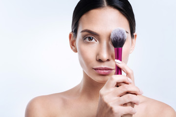 Pleasant dark-haired woman covering her eye with makeup brush