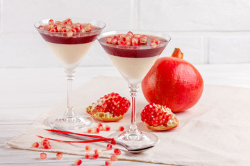Creamy vanilla panna cotta with red jelly in beautiful glasses, fresh ripe pomegranate on white wooden background.