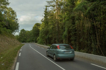 Cars driving on the asphalt road passing through the green forest in the region of Normandy, France. Summer nature, countryside landscape, transportation and road network concept.