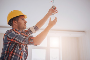 Handsome young builder in a yellow construction helmet is twisting the light bulb in. The man is looking up .