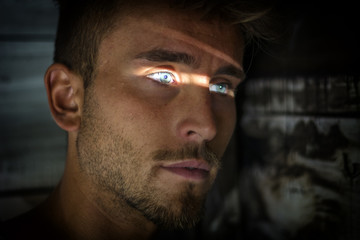 Handsome man leaning against wood planks wall outdoor with slit of light illuminating his blue eyes