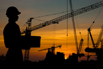 architect and engineer concept, Silhouette of working man standing and holding safety helmet and sheet of planning paper, building structure with crane and building background in sunset.