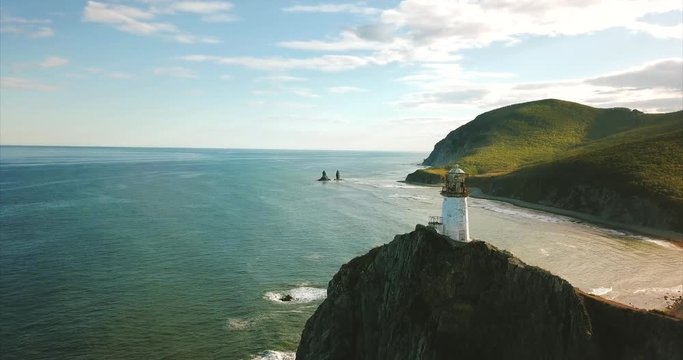 Slowly flying near the rocky cliff with lighthouse Brinera on it. It's an octahedral tower in the shape of truncated pyramid with lantern. Amazing seashore is on the background. Russia. Aerial