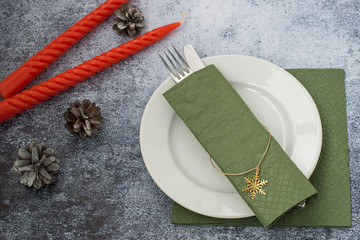     Preparing for a festive dinner. A round white plate, a paper green napkin, a fork and a knife on a dark blue background, next to red twisted candles and pine cones. © elizaveta66