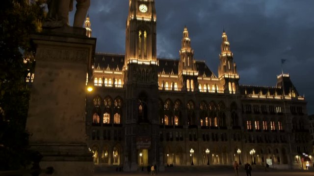 Tilt shot at night up the lit up facade of Rathhaus Vienna. Dark blue clouds cross the sky and framed by a statue