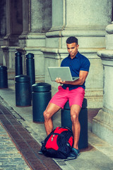 African American College Student traveling, studying in New York, wearing blue short sleeve shirt, red shorts, with backpack on ground, sitting on pillar on street, working on laptop computer..