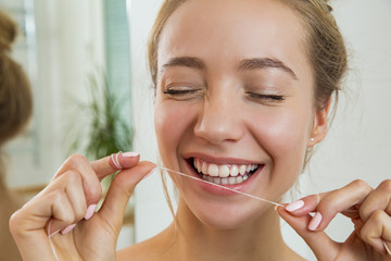 Young beautiful woman cleaning her teeth with floss in bathroom. Standing in towel, looking in the mirror, laughing and having fun. Daily routine. Beautiful smile with white teeth.