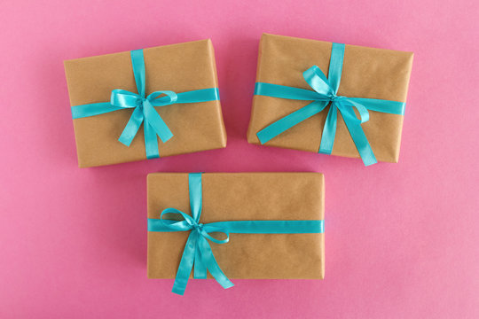 Three gift boxes wrapped of craft paper and blue ribbon on the pink background, top view.