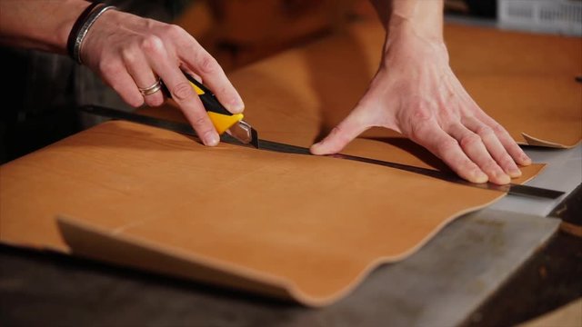 Man is carving details from treated brown skin. Master is holding main piece and applying line and stationery knife, slow motion close-up