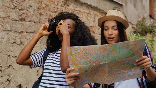 Two beautiful young multhi-ethnic girls holding camera and city map in hands looking attractions of the city in old street. Sightseeing. Outdoors.