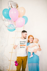 a young family of three people, mom's dad and daughter's one year old stands inside the room. Holding a balloon in her hand, a woman with red long hair is holding a child in her arms . Birthday party