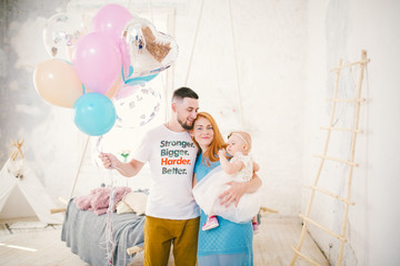 a young family of three people, mom's dad and daughter's one year old stands inside the room. Holding a balloon in her hand, a woman with red long hair is holding a child in her arms . Birthday party