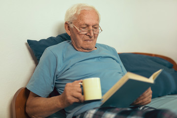 Senior man lying on bad and reading book at home