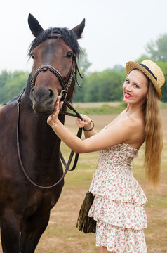 Smiling girl in a straw hat and summer dress, photographed with a horse / Photo taken in Russia, at the hippodrome of the city of Orenburg