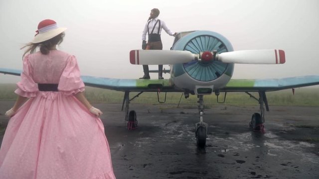 A woman in a pink dress of 60th comes to a vintage airplane on which stands the pilot
