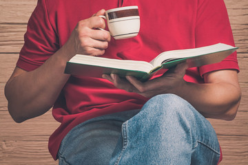 a young man in a red t-shirt and blue jeans in one hand holds a book, and in another cup.