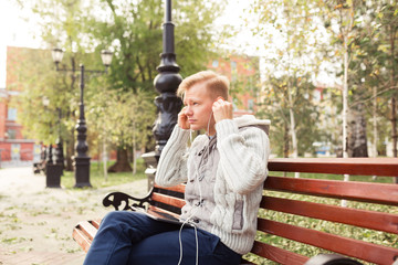 Handsome young man with white headphones sits on a bench in the city at autumn