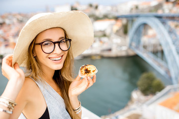 Portrait of a young woman enjoying traditional portuguese dessert called pastel de Nata, sitting on the terrace with beautiful cityscape view on Porto, Portugal