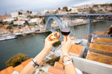 Holding a glass of red wine with traditional portuguese dessert called pastel de Nata on the beautiful cityscape background in Porto, Portugal - obrazy, fototapety, plakaty