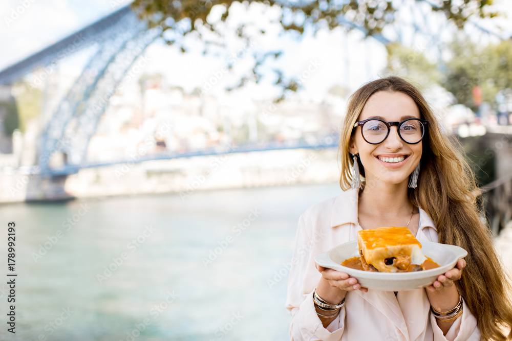 Wall mural Portrait of a young woman with traditional portuguese sandwich with meat called francesinha on the landscape background with bridge in Porto city, Portugal