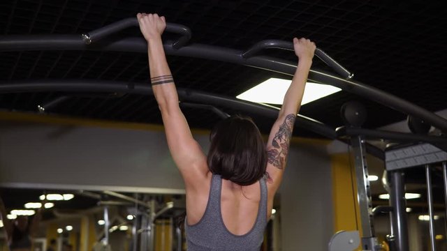 Athletic woman with tattoos is doing an exercise for her back in gym. She is doing clear pullups.