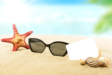 Vacation concept image - sunglasses, shells, starfish and blank white card on a sandy tropical beach in close-up and on a blurred coastline background.