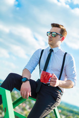 Fototapeta premium Attractive young busunessman in white shirt, tie, braces and sunglasses with red cup
