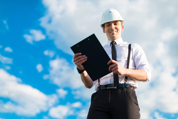 Young attractive foreman in white helmet, tie and braces stand on the roof with tablet