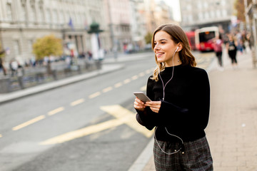 Fototapeta premium Young brunette woman using mobile while standing on street and waiting for a bus or taxi