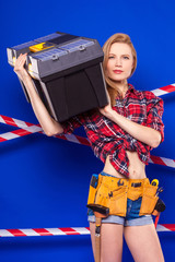 Young slim builder girl in red chechered shirt, builder belt, jean shorts and snickers hold toolbox