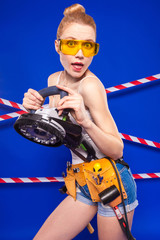 Pretty, slim builder girl in white shirt, builder belt, builder glasses, jean shorts and snickers hold electro saw