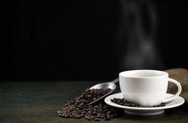 Hot coffee in the white cup with roast coffee beans, bag and scoop on stone table in black background