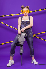 Beauty young builder girl in denim jumpsuit and glasses stand with puncher