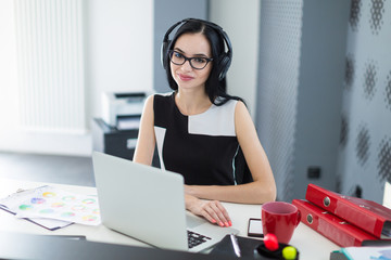 Beautiful young businesswoman in black dress, headphones and glasses sit at the table and work on laptop