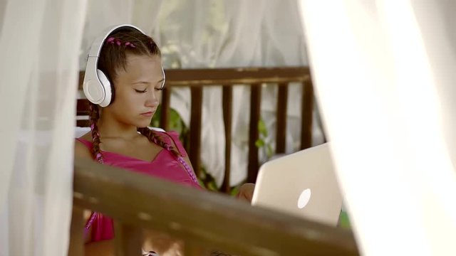 teenage girl is hearing music by headphones, sitting on a terrace in sunny day, holding open laptop