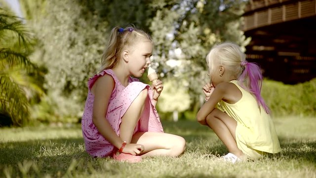two little girls with blond hair are sitting on grass in a yard in countryside in sunny summer day