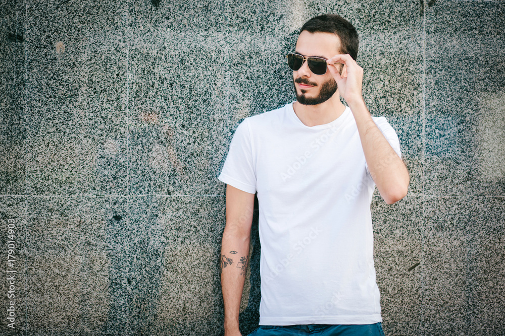 Sticker Young man wearing white blank t-shirt with beard in glasses, standing on the street on city background. Street photo