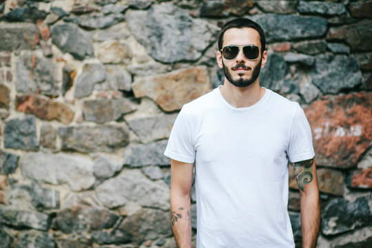 Young man wearing white blank t-shirt with beard in glasses, standing on the street on city background. Street photo