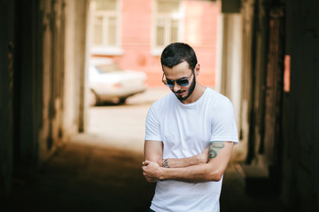 Young man wearing white blank t-shirt with beard in glasses, standing on the street on city background. Street photo