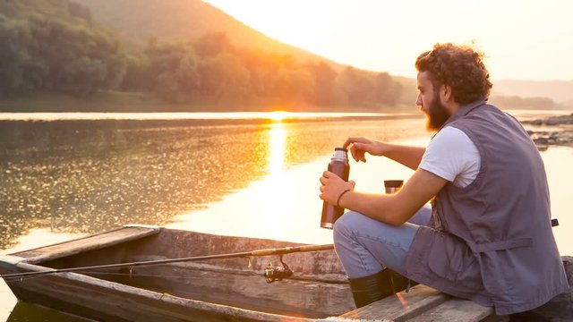 Stylish hipster with thermos at sunset. Fisherman in an old wooden boat on a summer evening